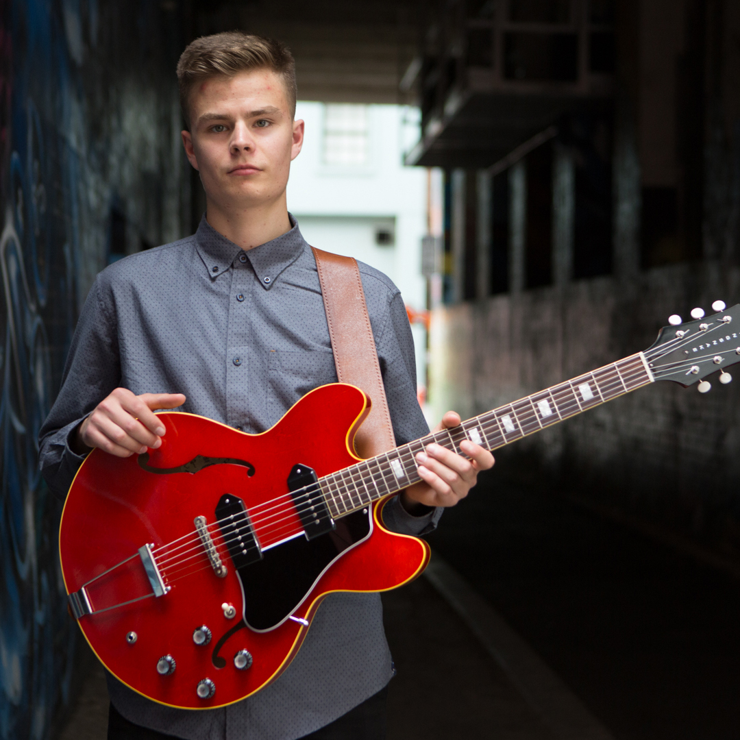 A young, white male holds a red guitar and faces the camera. He wears a grey shirt with a collar and s standing in a alleyway