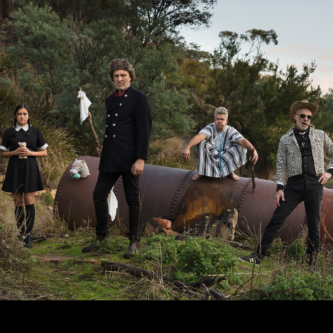 A group pf people stand in the bushland. The man at the front wears a russian hat and holds a white flag on a stick. The man on the left wears a cowboy hat and a clerical collar. A man in the background wears a mumu and has his eyes closed and looks to be holding his breath with his cheeks puffed out. The woman on the left wear a black dress with a white collar and holds a teddy bear.