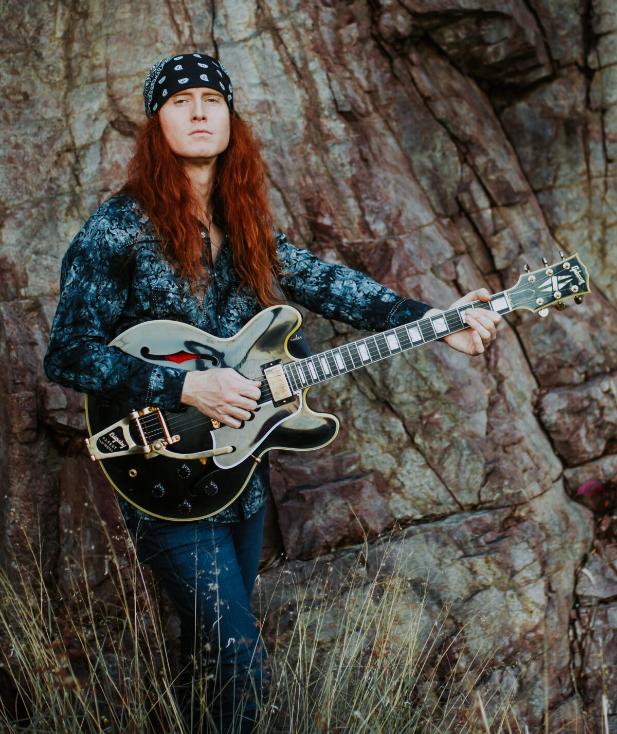 A white man with long red hair stands in front of a tree with a black guitar. He wears a black bandana on his head.
