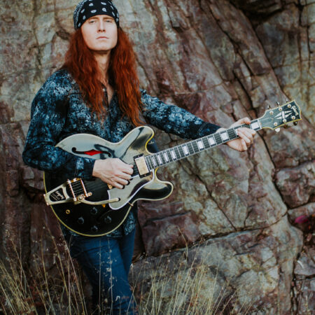 A white man with long red hair stands in front of a tree with a black guitar. He wears a black bandana on his head.