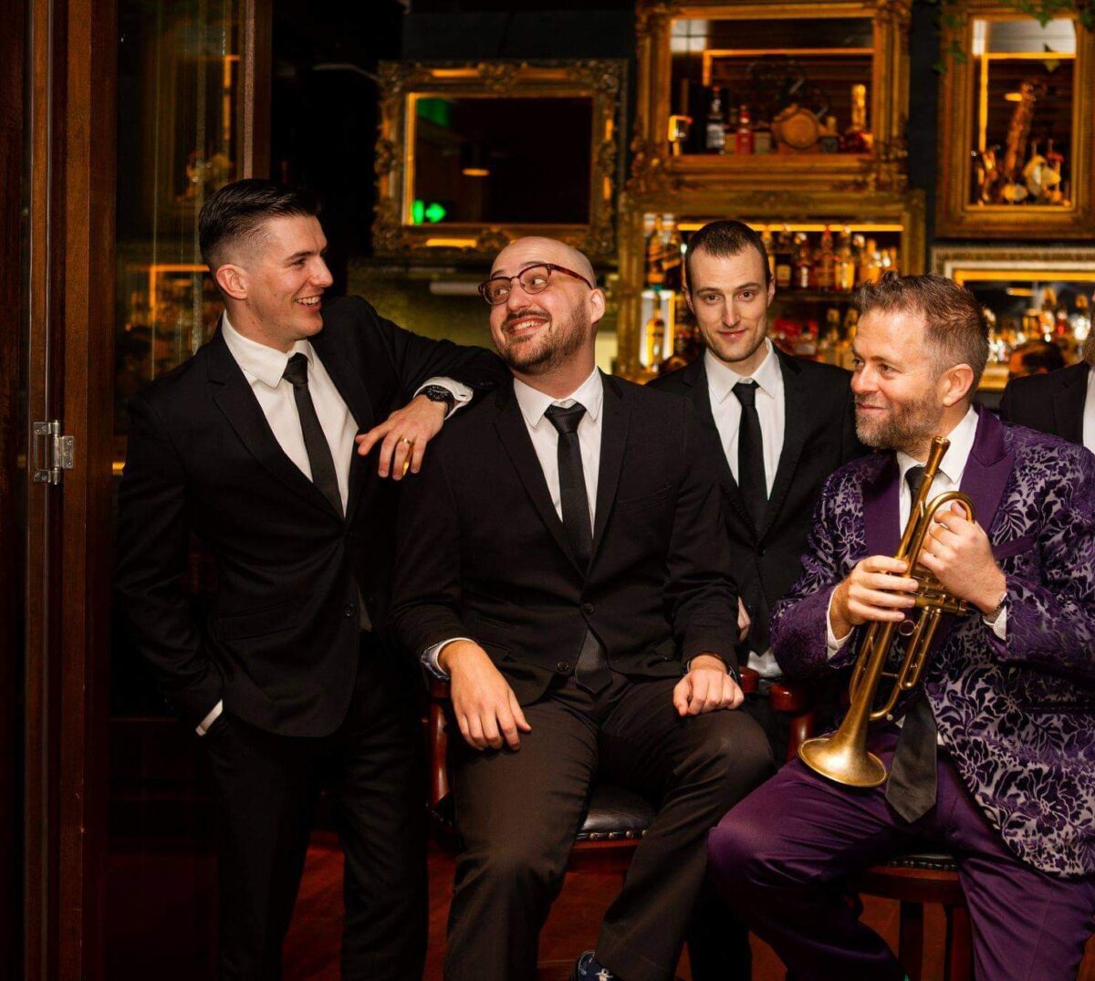 A group of white men in suits sit in a bar. One of them holds a trumpet.
