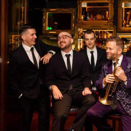 A group of white men in suits sit in a bar. One of them holds a trumpet.