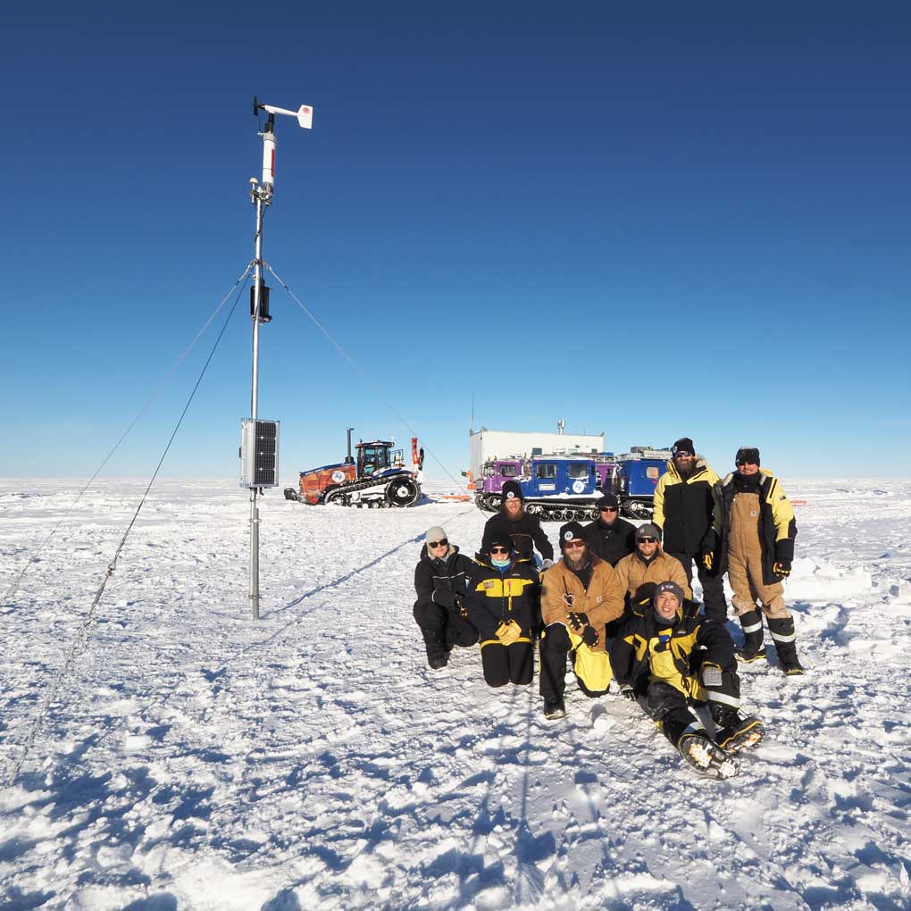 A photo of a group of people dressed in thick coats and hats in Antarctica. They are sitting on the ice, next to a weather pole, and in the background there are sevral vehicles for travelling in the ice and snow.