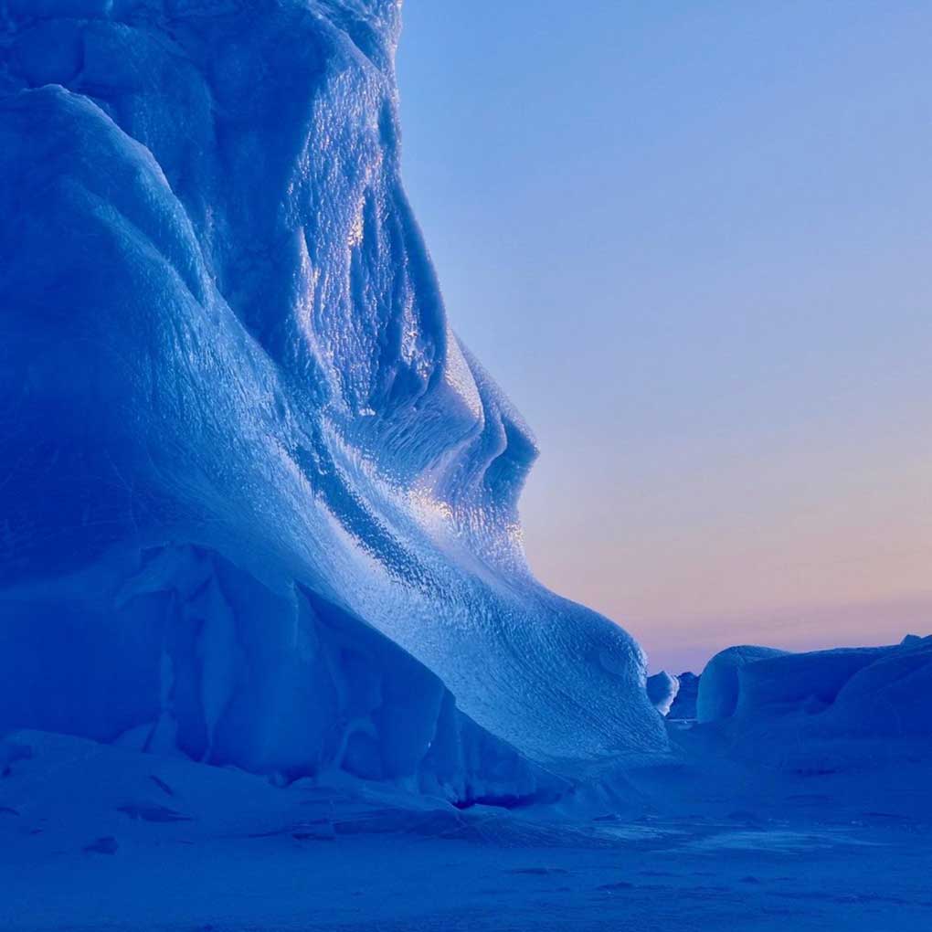 Photo of a large ice cliff in Antarctica. The cliff cliff of ice is a deep blue and rises to the right of the photo. The sky in the background is pastel tones, indicating dawn or sunset.
