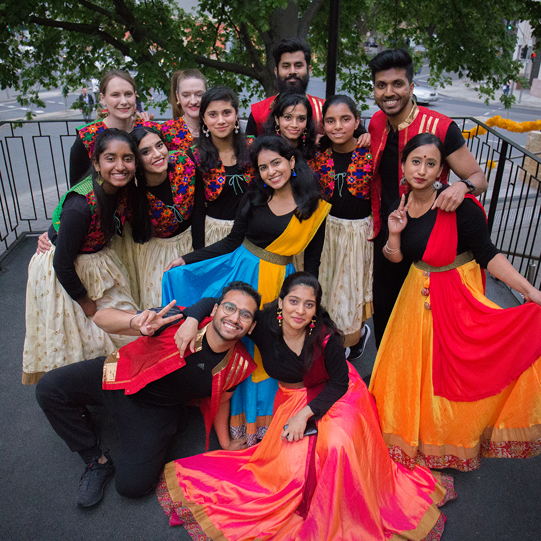 A group of colourfully dressed Indian dancers are grouped together. They are smiling and looking at the camera.