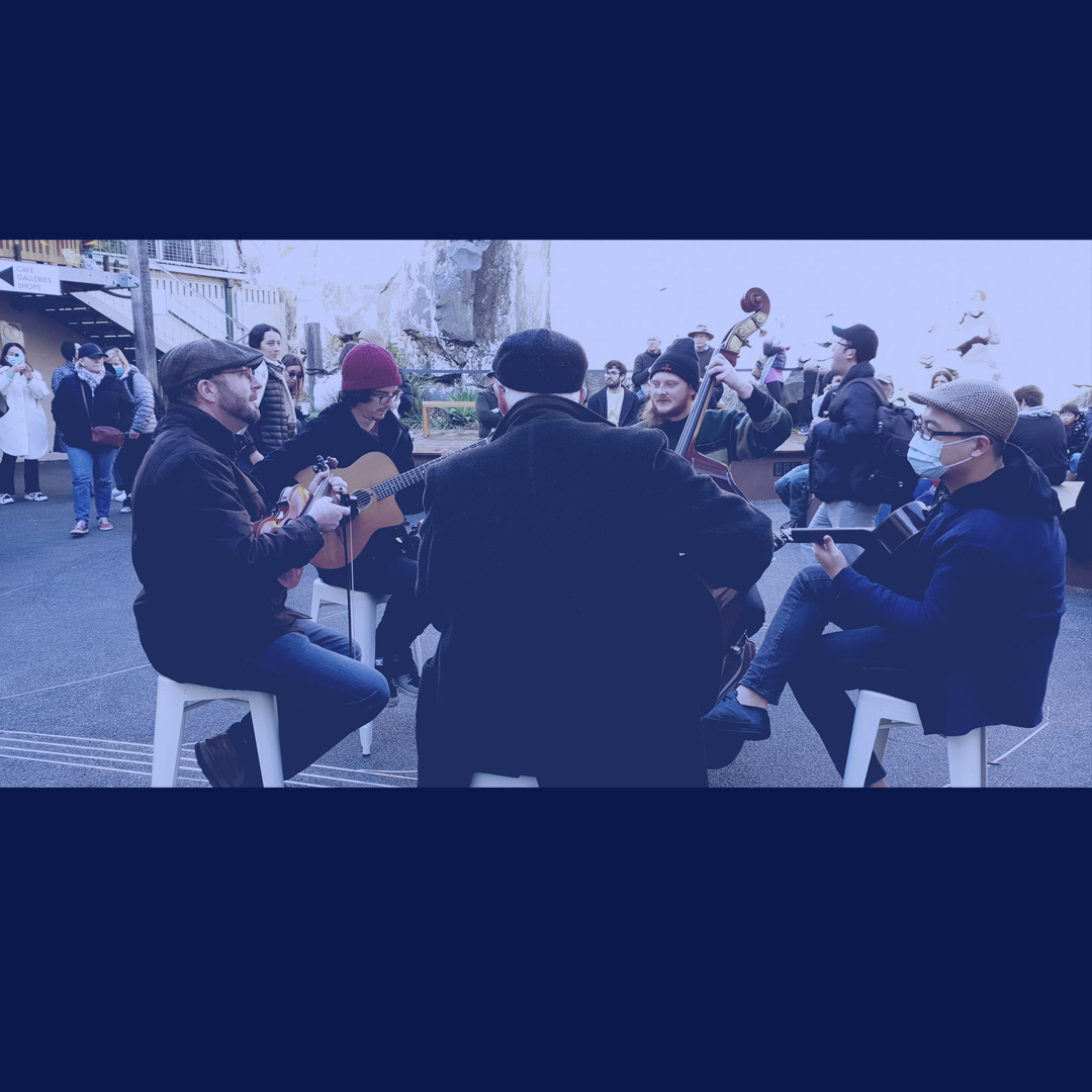 A group of musicians play in a circle. They are all male and white. They play in an outdoor area.