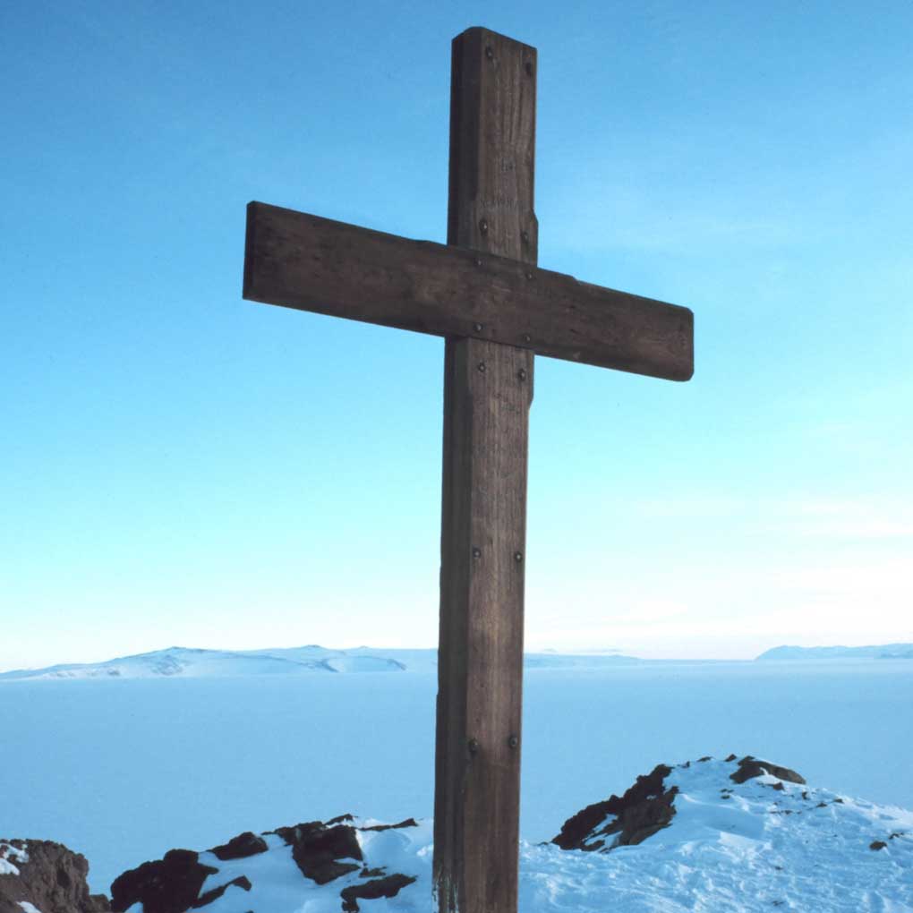 A photo of a dark wooden cross in the left hand foreground, perched on a rocky outcrop covered in snow and ice.