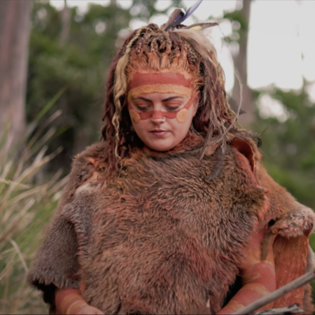 A palawa woman wears a fur cloak and has ochre on her face. She looks downward and has long hair.