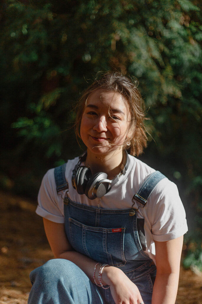 A woman wearing headphones around her neck looks directly to camera. She is in the sunshine and has her brown hair pulled back in a ponytail. 