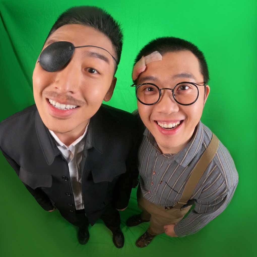 Two actors photographed against a green background at a very high angle; their faces are closer to the camera. One actor is dressed in a black suit and is wearing an eye patch. The other actor is dressed in a stripe shift with suspenders, and is wearing glasses; he has a bandage on the side of his head.