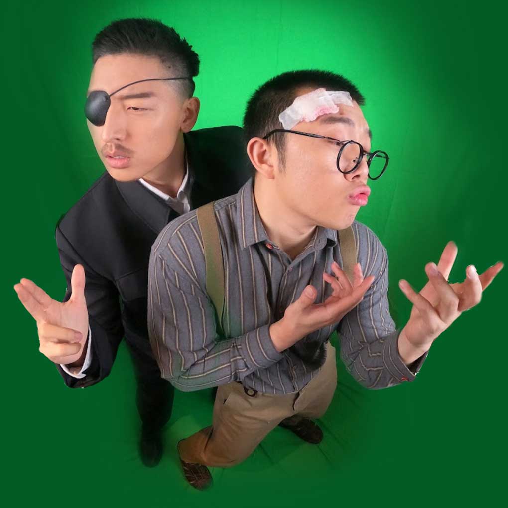 Two actors photographed against a green background at a very high angle; their faces are closer to the camera. Both actors are posed dramatically.One actor is dressed in a black suit and is wearing an eye patch. The other actor is dressed in a stripe shift with suspenders, and is wearing glasses; he has a bandage on the side of his head.