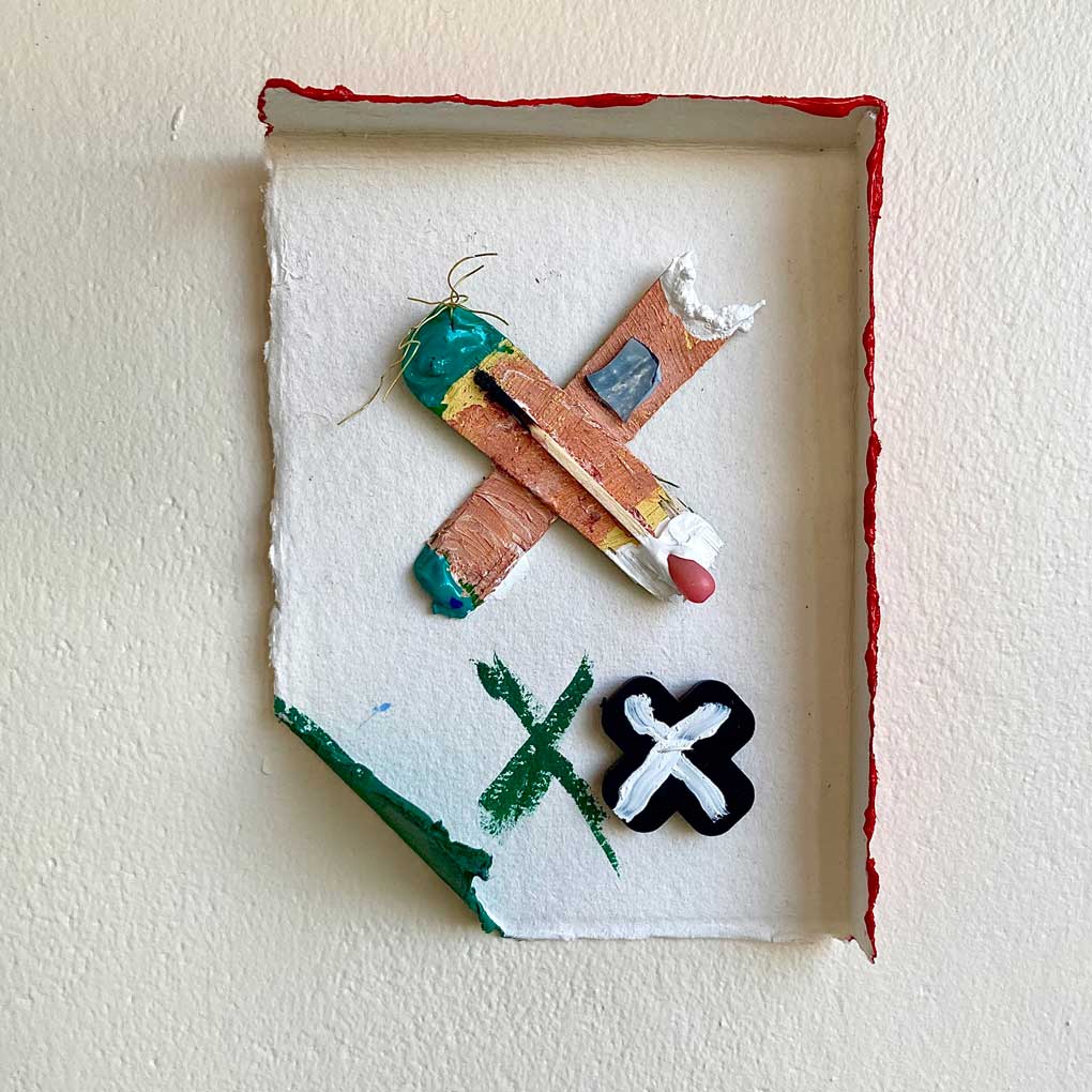 A small mixed media assemblage against a white background. Within a white paper frame there are three crosses, one painted green, one painted black with a white line ontop, and the third created from scraps of brown and green paint, topped with a matchstick.