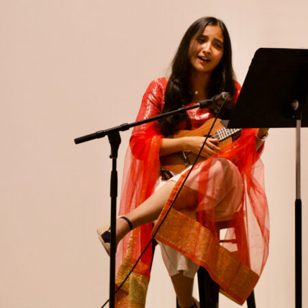 An indian woman with long dark hair sits on a tall stool wearing orange. A microphone and music stand is in front of her.