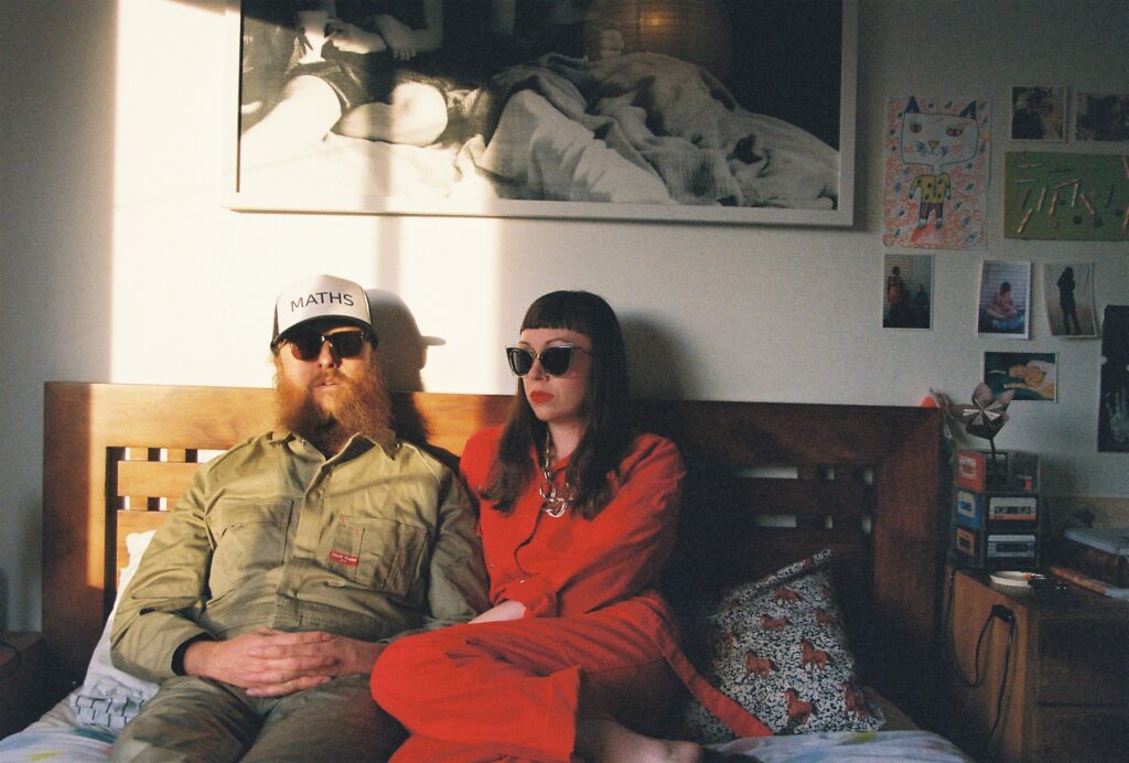 A Couple sit on a lounge. She wears a red street and sunglasses and has long dark hair. He wears a cap, sunglasses and a long ginger beard. 
