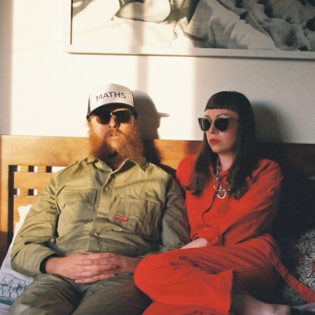 A couple sit on a lounge. She wears a red street and sunglasses and has long dark hair. He wears a cap, sunglasses and a long ginger beard.