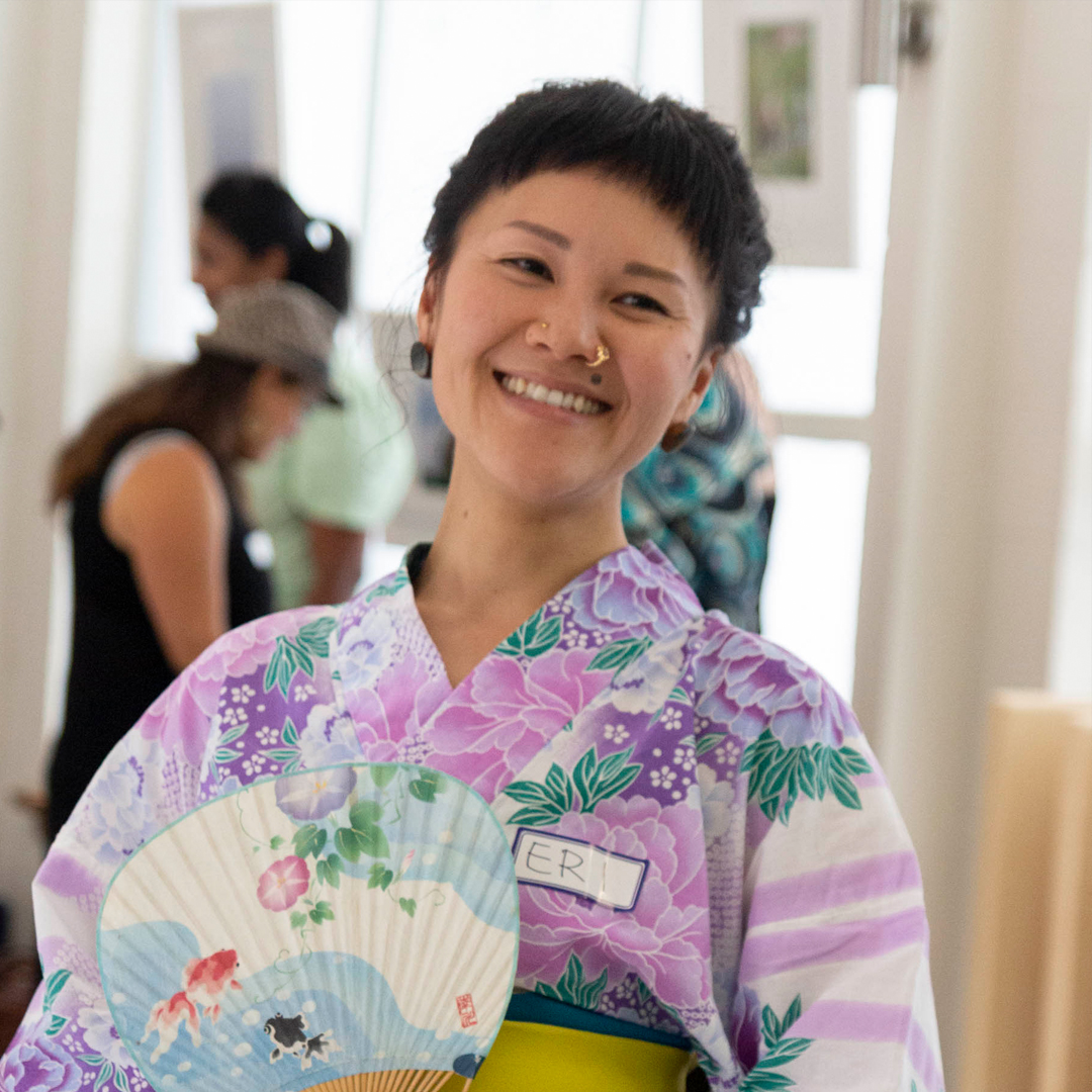 A japanese woman with a dark fringe and smiley face looks directly to camera. She wear traditional Japanese clothing in pastel purple, pink and white and holds a traditional fan.