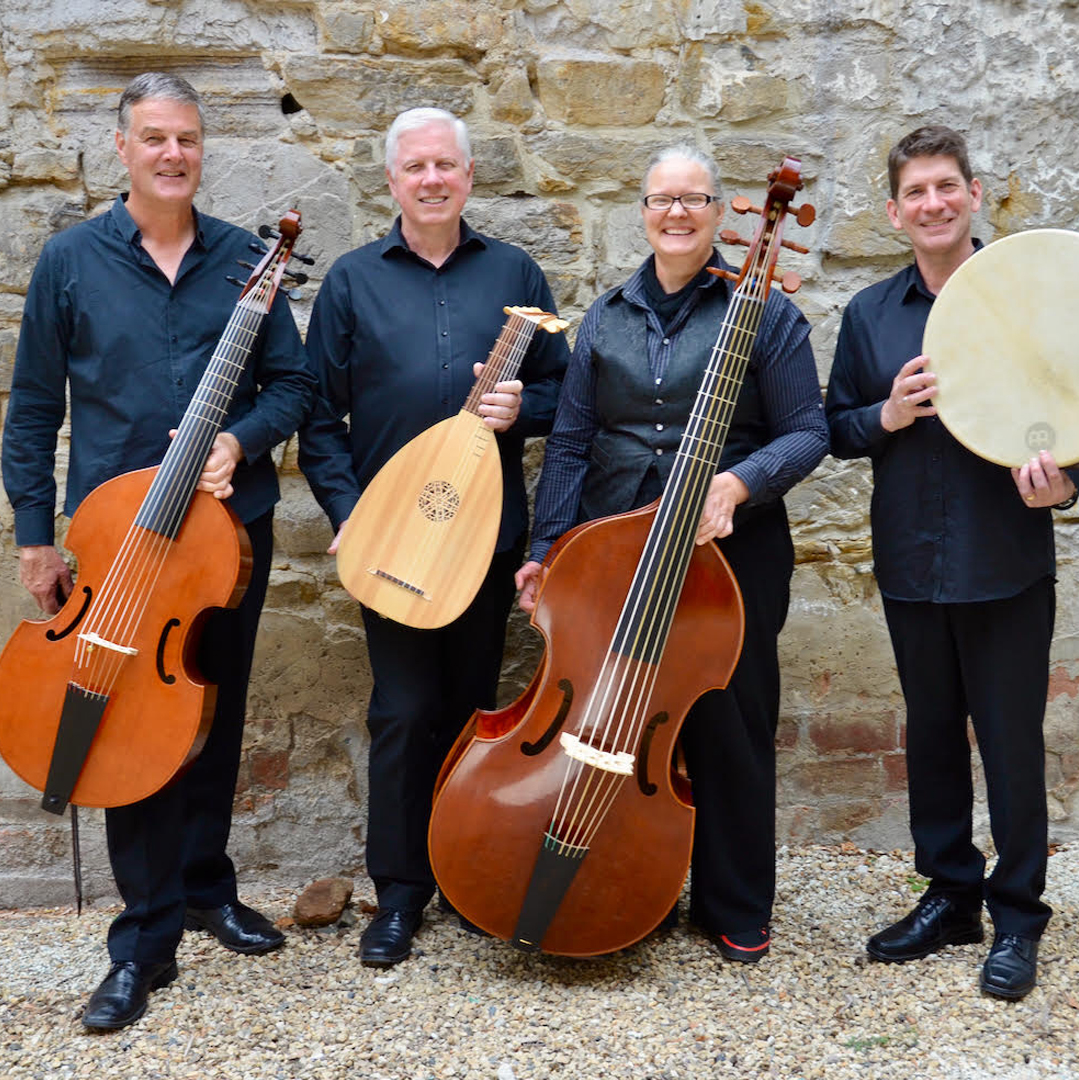 Four people stand in front of a convict sandstone wall. They each hold an instrument and are wearing black clothing. They hold a cello, lute, double bass and drum. They are all white and three of them have grey hair. One has brown hair. They all look very happy and are looking directly to camera.