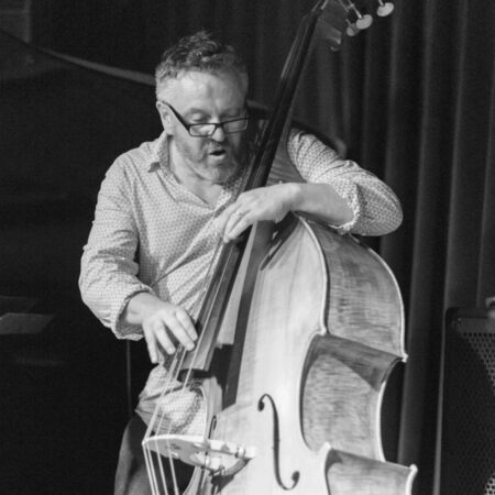 A black and white image of a white man playing a double bass. He wears glasses and a collared shirt with the top button undone.