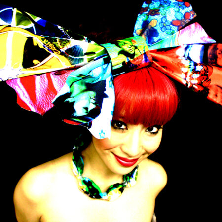 A photo of a Japanese woman from the shoulders up. She has a large colourful bow in her hair and has a red coloured fringe.
