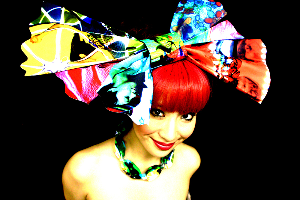 A photo of a Japanese woman from the shoulders up. She has a large colourful bow in her hair and has a red coloured fringe.