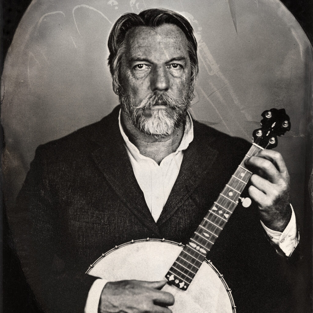 Black and White tintype photograph of a man holding a banjo. The man is wearing a dark jacket and white shirt, and has a beard.
