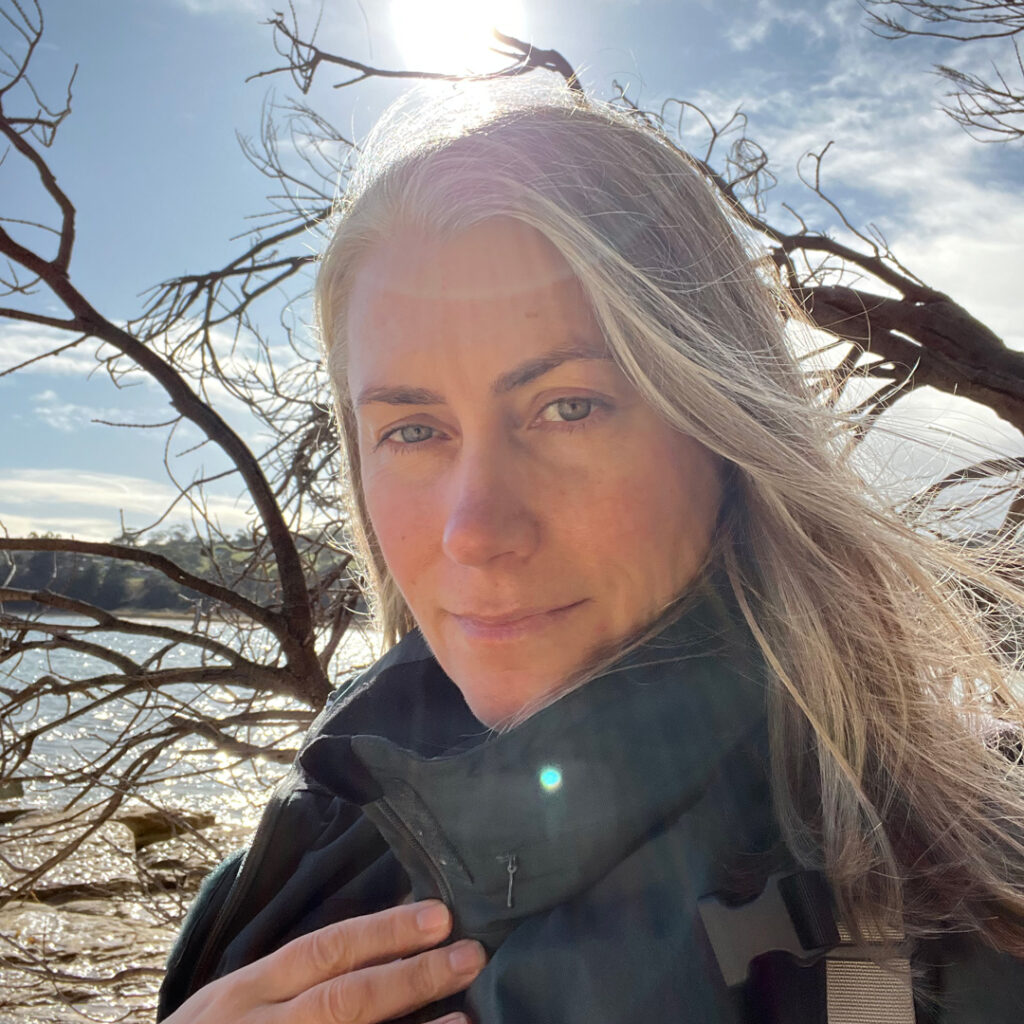A photograph of woman with blond hair standing on the foreshore. In the background are trees with twiated branches.