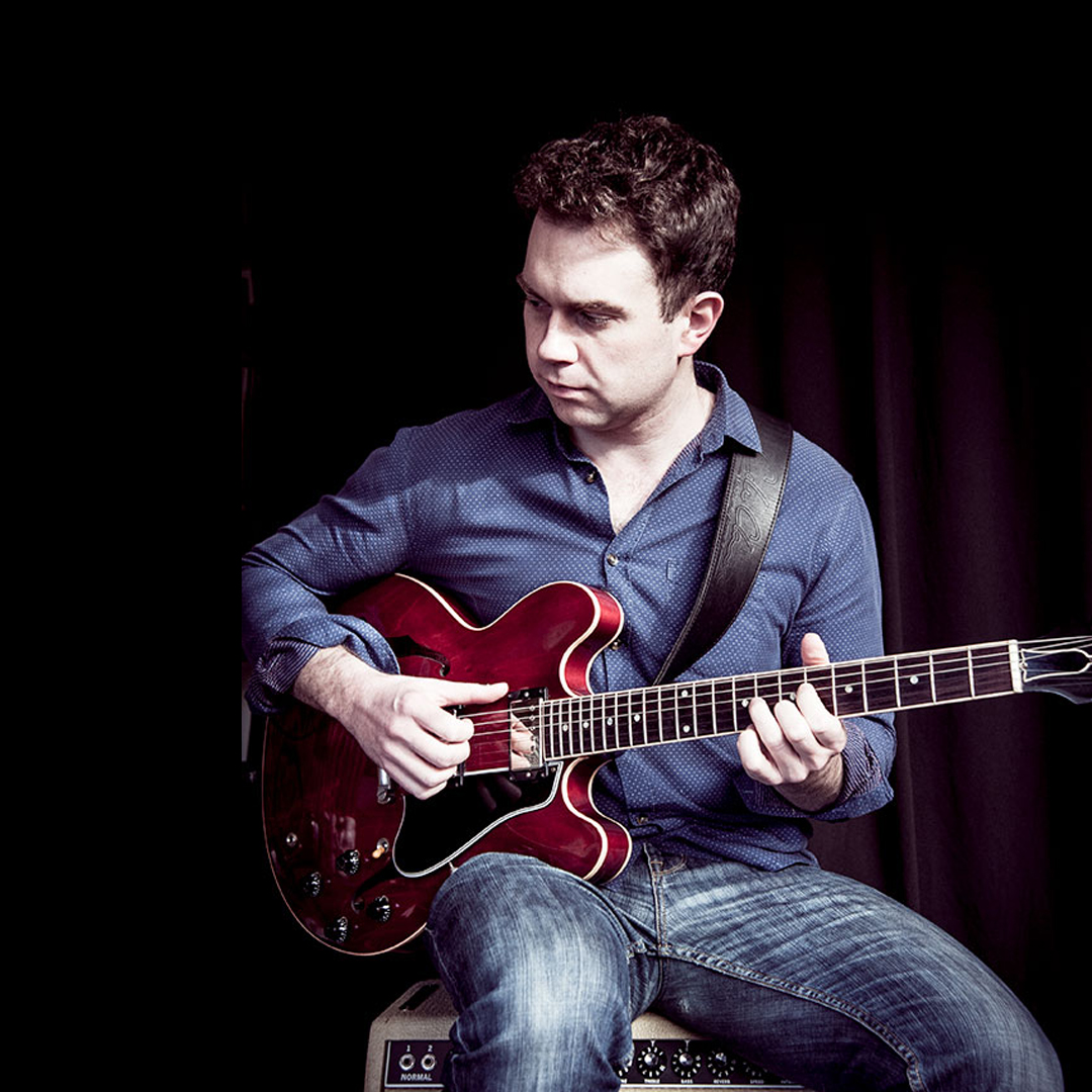 A white male sits on a seat holding a red guitar. He looks to the left of the image.