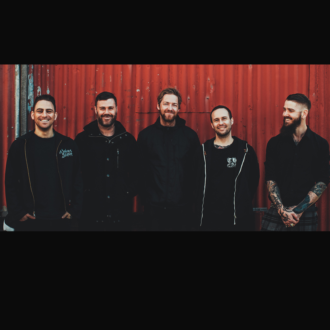 A group of 5 men stand in front of a red corrugated fence. They are all wearing black and all but one are looking directly to camera.