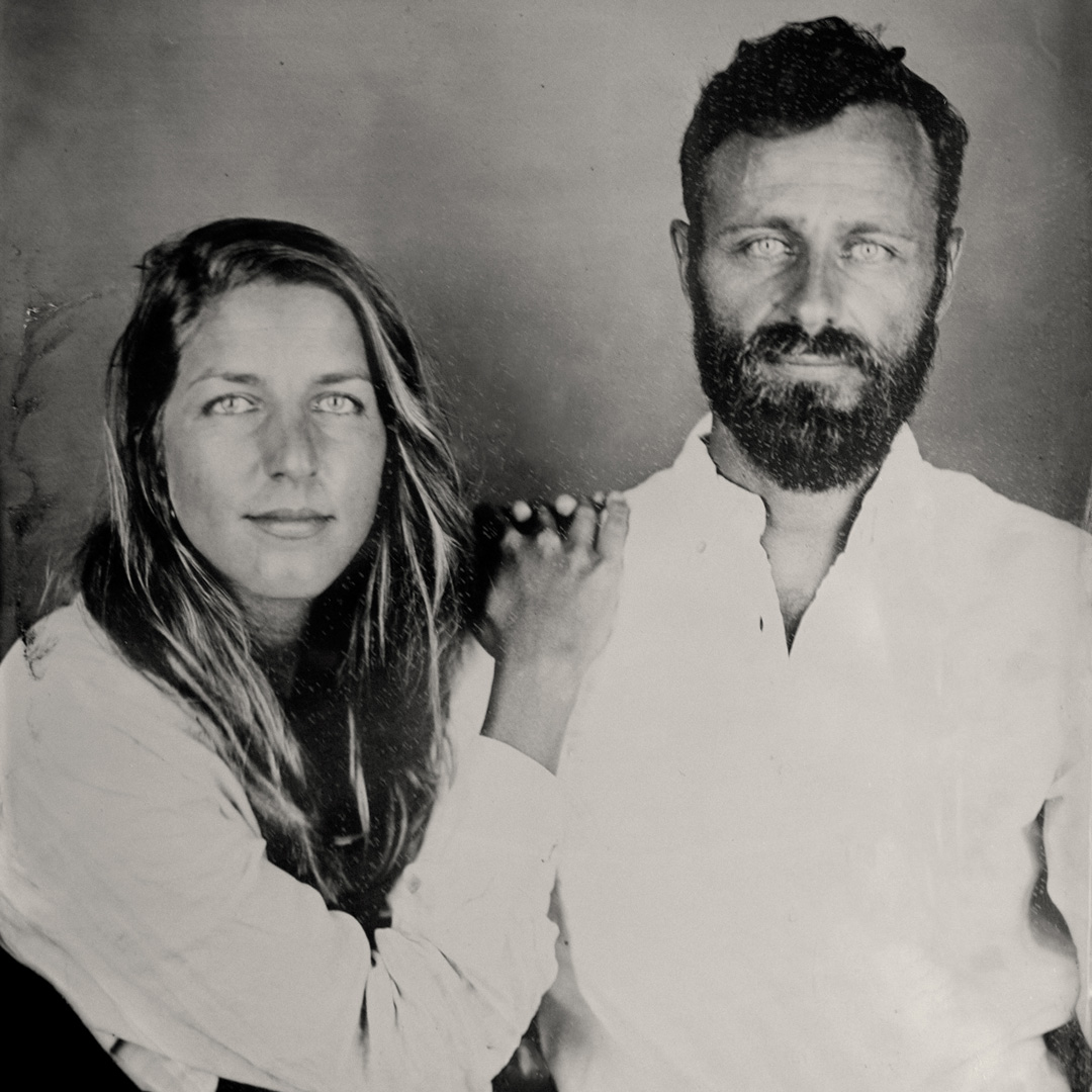Black and White tintype photograph of a woman with long hair leaning against the shoulder of a man with dark hair and a breard. Both are wearing white long-sleeved shirts.