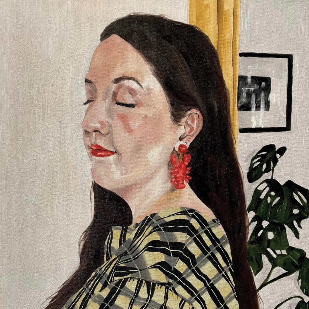 Portrait of a lady with long, dark hair with bright red lipstick and red lobster earrings. She is wearing a yellow and black checked top, and in the background there is a yellow curtain, a black and white framed photograph hanging on the wall and the leaves of an indoor cheese plant.