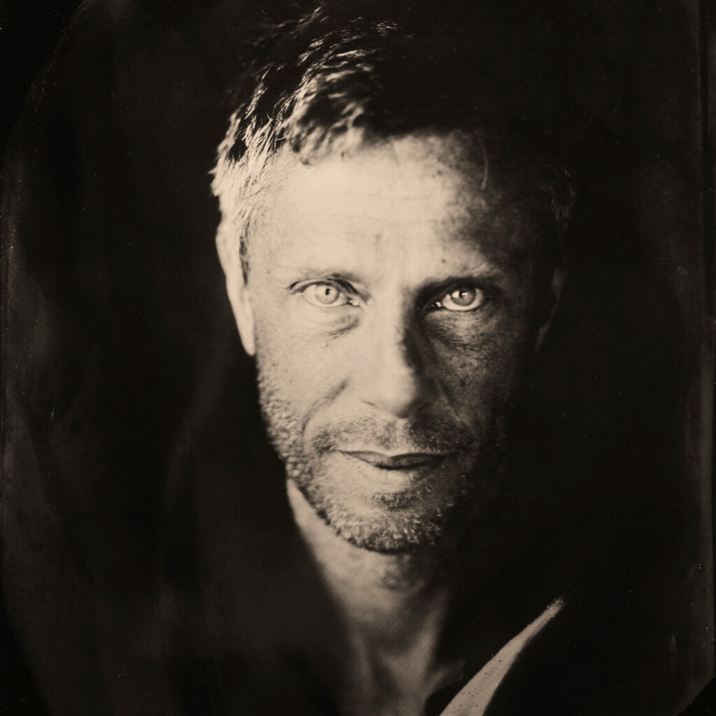 Black and White tintype photograph of a man's face. The backgroud is very dark and the only light is on the left side of the face.