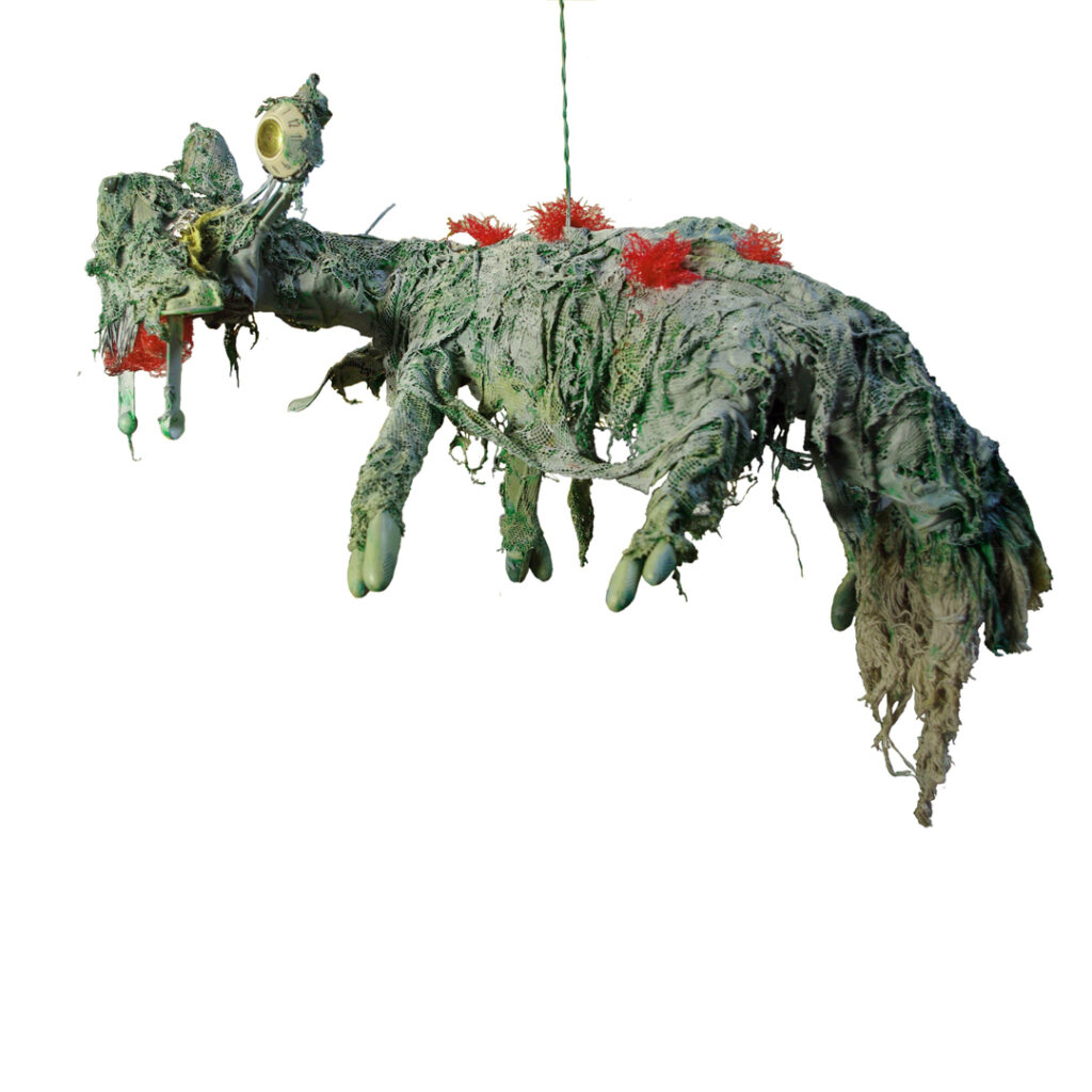 A green creature suspended from a thin cord. The creature is made from found objects, including garbage ags and rubber gloves and has tufts of red sprouting from it's back.
