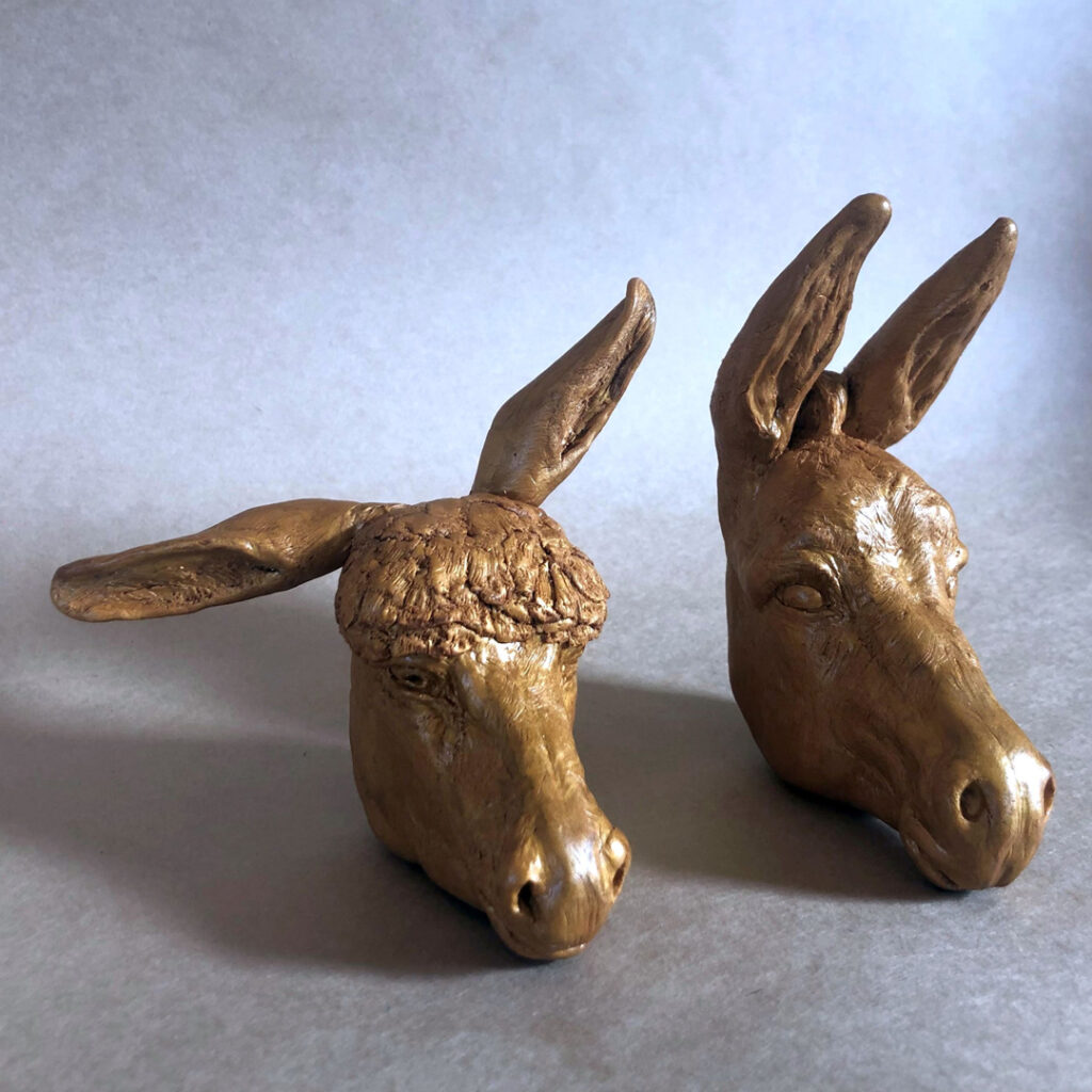 Two ceramic donkey's heads. The head on the left has curly hair on it's head and ears twisted to the side. Whilst the other donkey's ears are blowing in the wind.