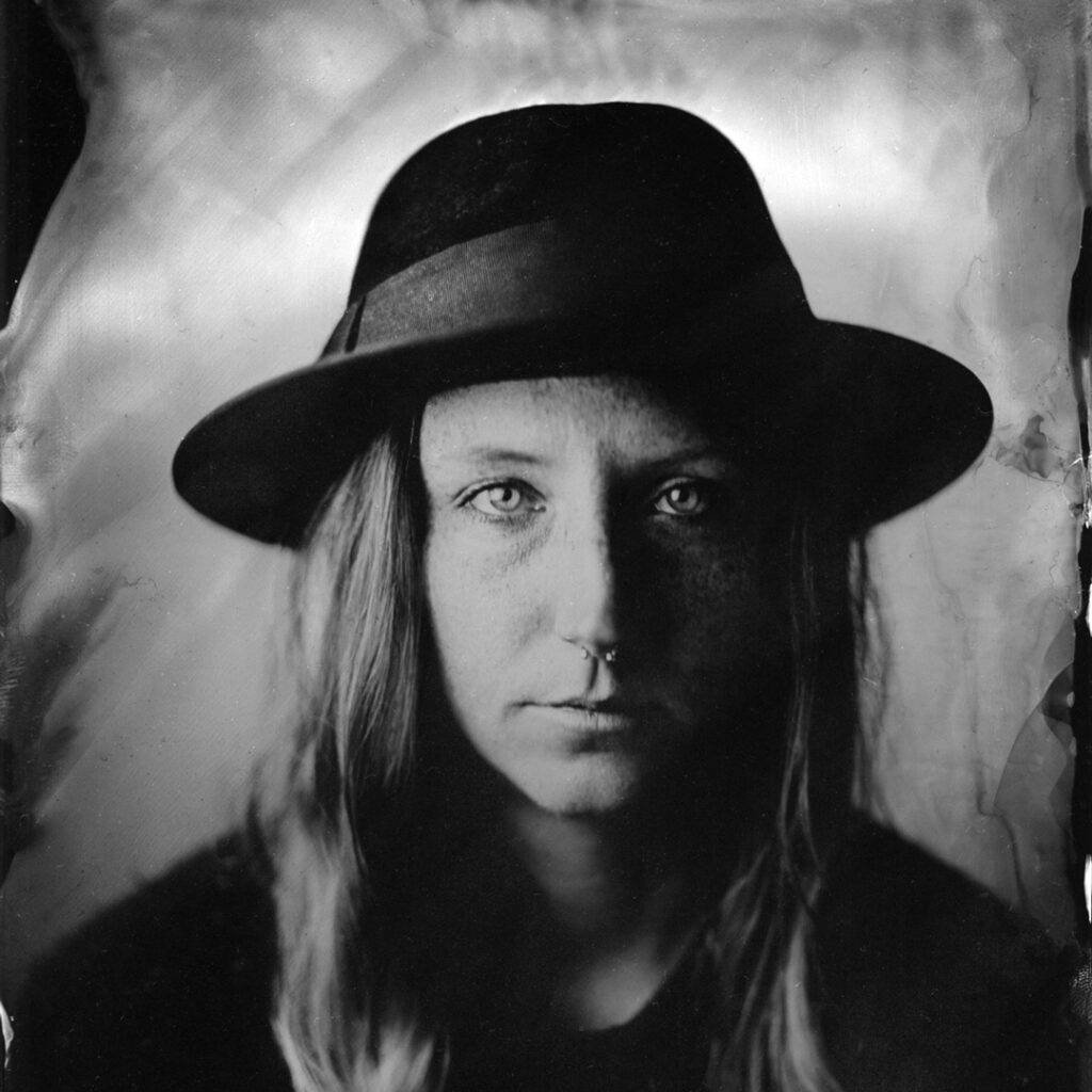 Black and White tintype photograph of a person with long hair wearing a black fedora hat.