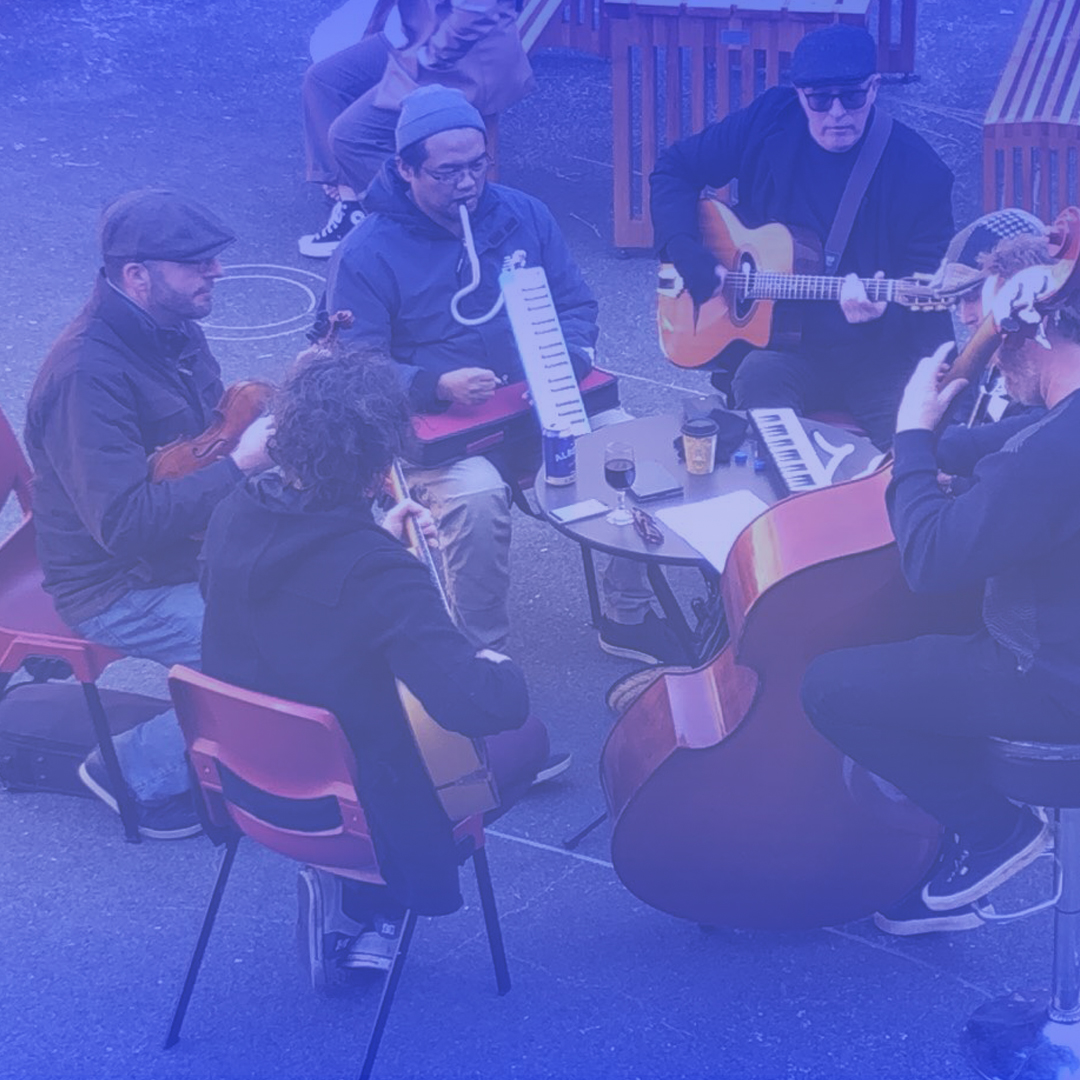 A group of musicians play in a circle. They are all male and white. They play in an outdoor area.