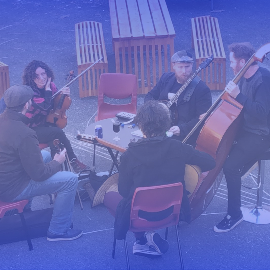 A group of musicians play in a circle. They are all male and white. They play in an outdoor area.