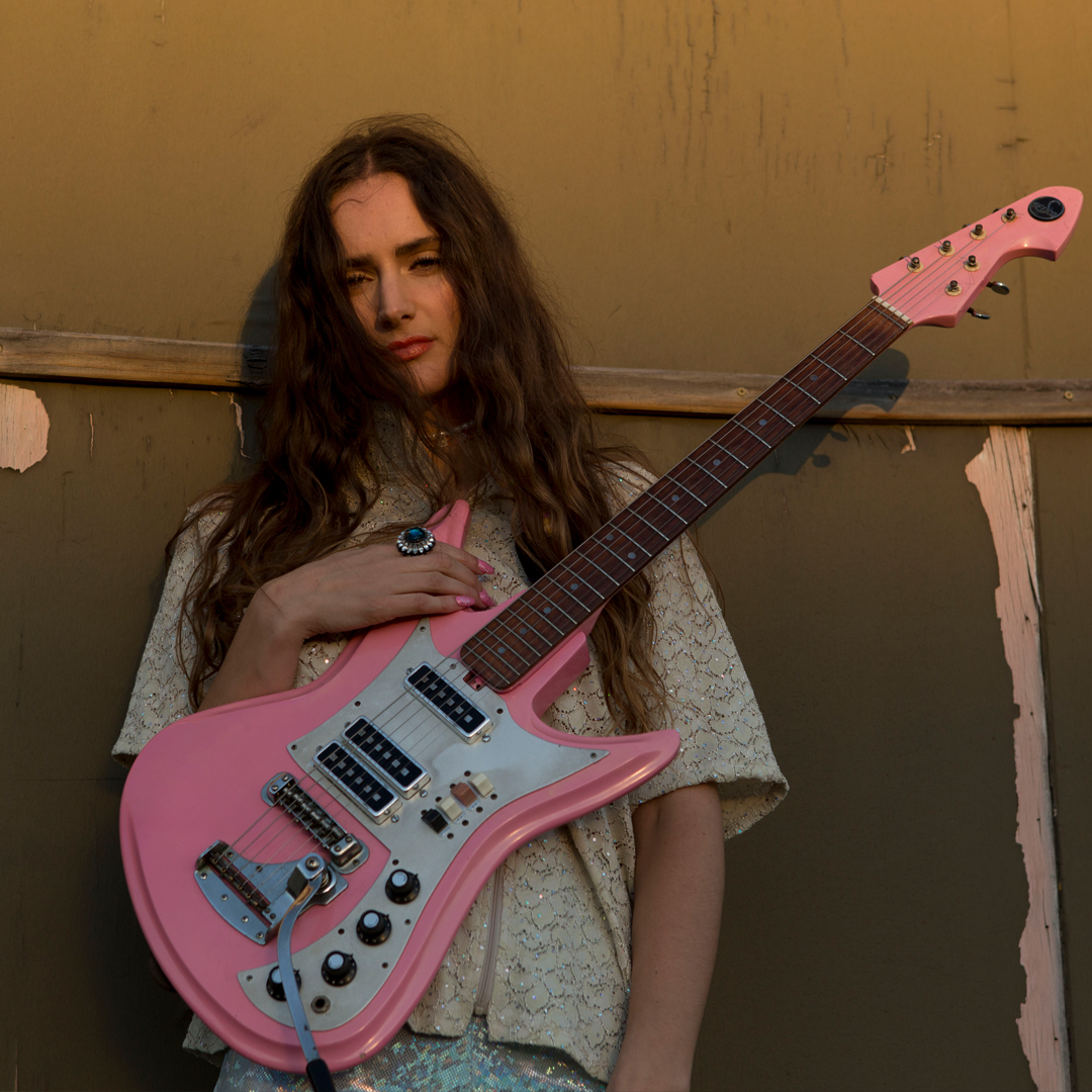A girl with long dark hair holds a pink guitar and looks directly to camera. She stands in front of a tan coloured wall with peeling paint.