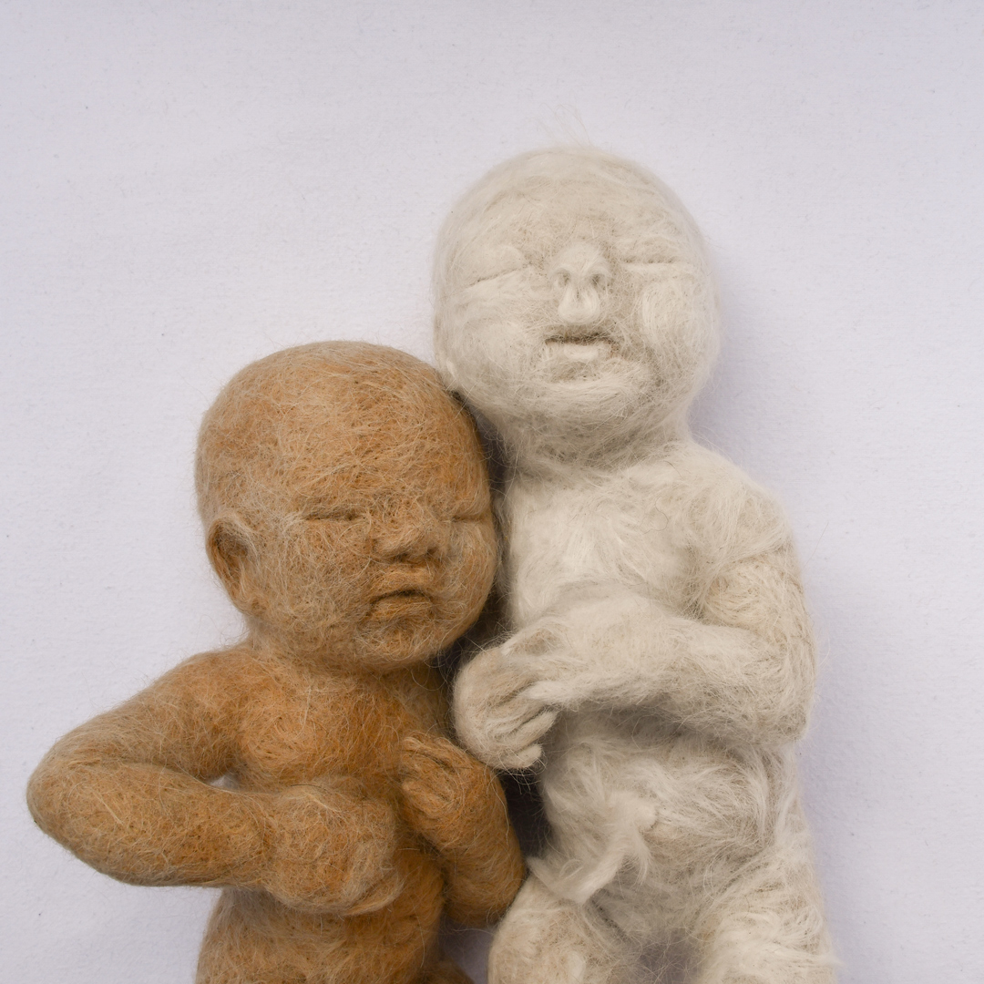 A human babies - one light orange brown and one white - created from felted dog hair, against a white background.