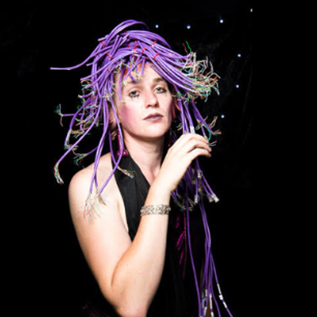A woman wearing a purple wig stands in front of a black, starry background. She looks directly to camera and holds a drink in her hand.