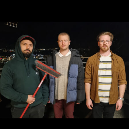 Three men look to camera. One of them holds broom.