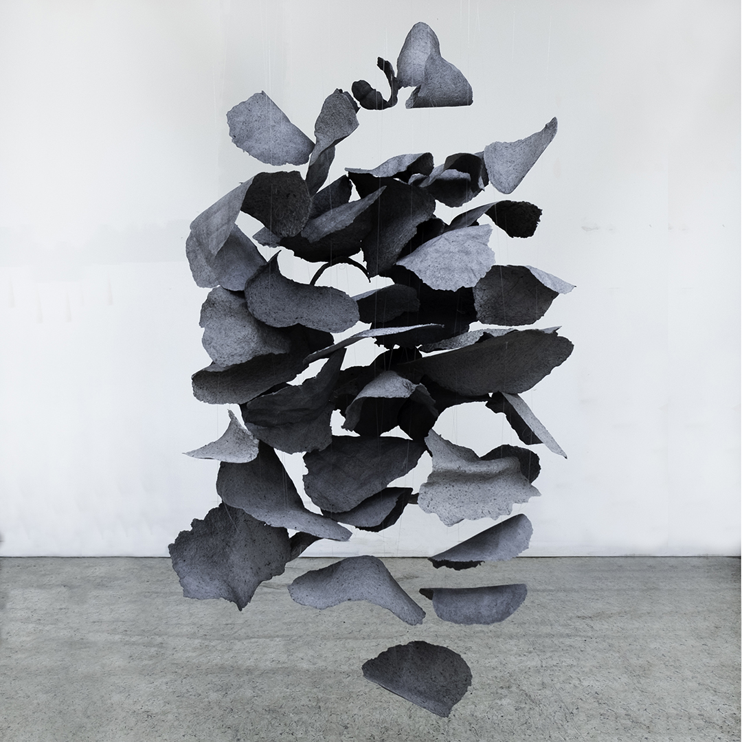 grey paper petals hover in the air in front of a white wall. The floor is concrete.