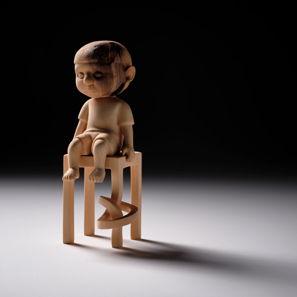 A wooden carving of a figure sitting on a chair. One leg of the chair is tied in a knot.