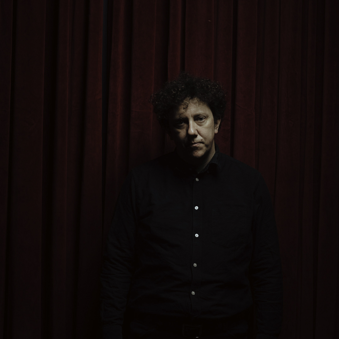 A white man with dark curly hair stands in front of a red velvet curtain. He looks directly to camera.