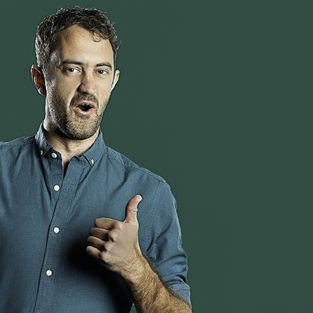 A white man wearing a green collared shirt stands in front of a green background giving a thumbs up sign.