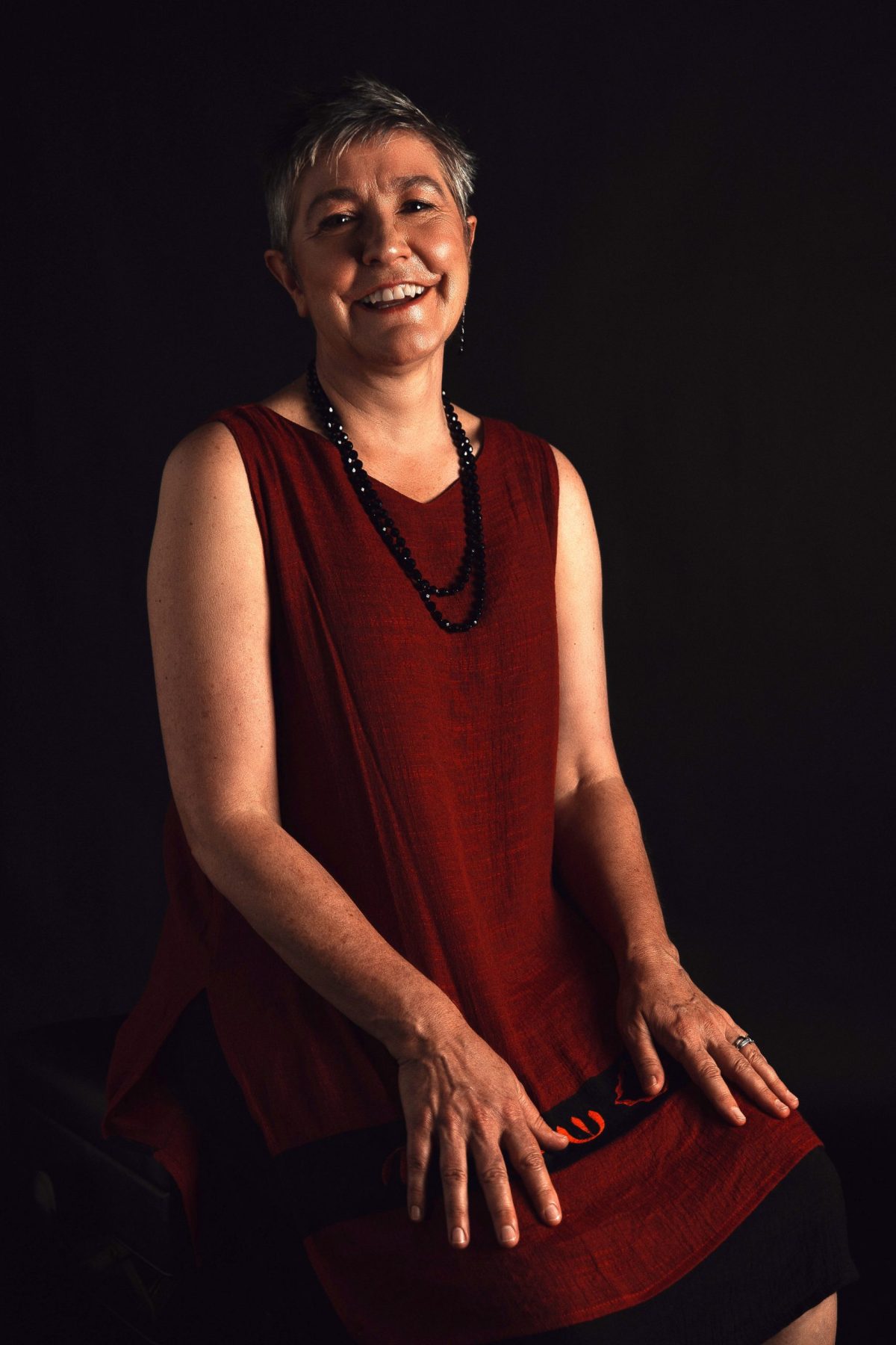 Smiling lady with glasses and grey hair in a maroon dress against a black background